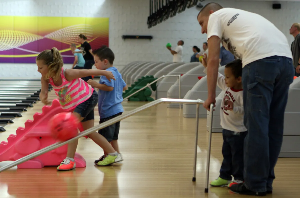 Children rolling a bowling ball down a ramp, with a parent beside them on the lane