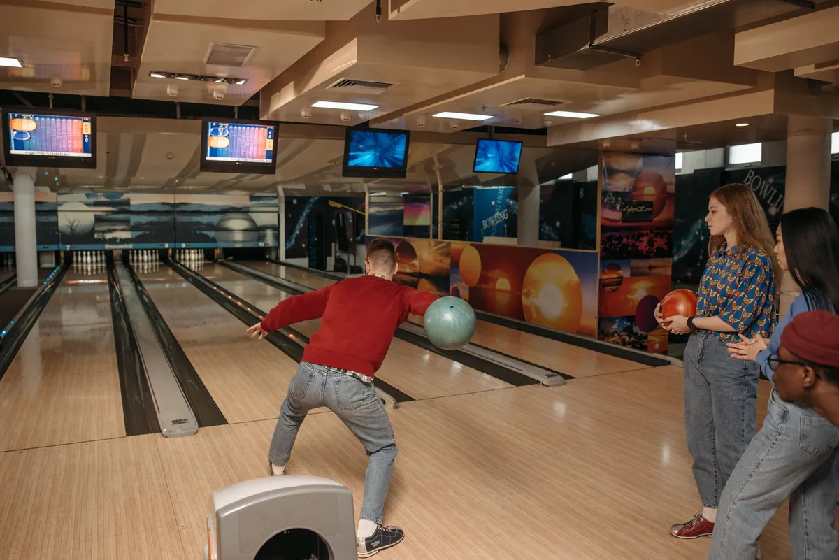 A bowler releasing the ball while friends wait patiently for their turn behind the approach
